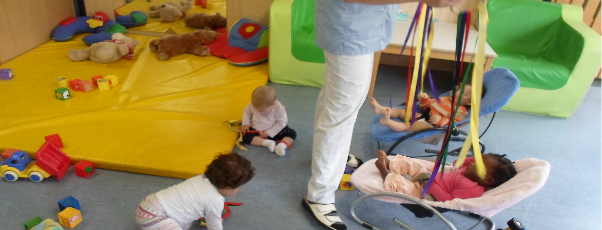 Deux enfants jouent au sol avec des bandelettes et deux autres enfants sont couchés dans un coufins et la puéricultrice leur montre les bandelettes