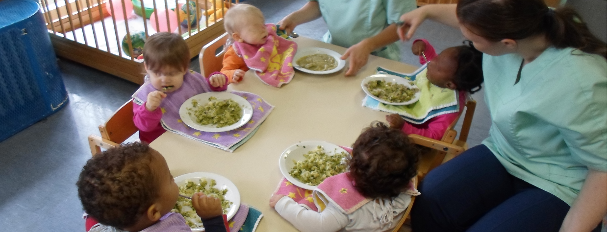 Des enfants mangent à table avec l'aide de deux puéricultrices