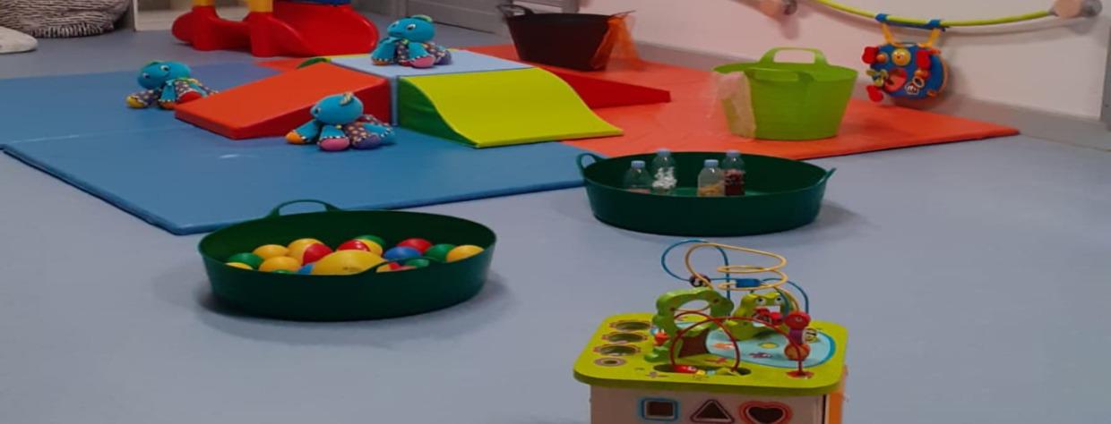 3 peluches e pieuvre posées sur des tapis en mousse avec deux bassines contenant des balles ou des bouteilles en plastiques. En avant plan il y  a un jeu de labirynthe